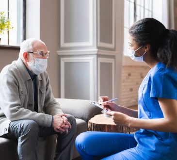 Young woman health aide consulting with older male patient during home visit.