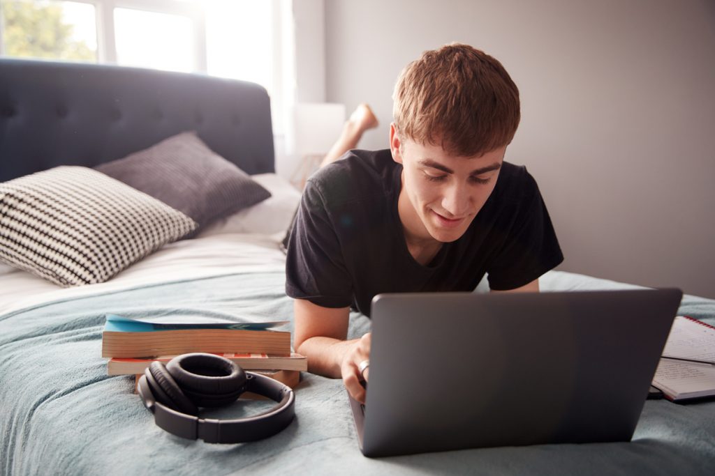 Male College Student Lies On Bed Working on Laptop