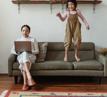 Woman working on laptop on couch while child jumps beside her.