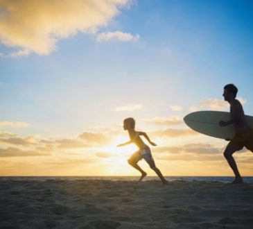 Adult and child silhouetted with surfboard on beach.