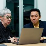 Older woman and younger male colleague talking over laptop in office