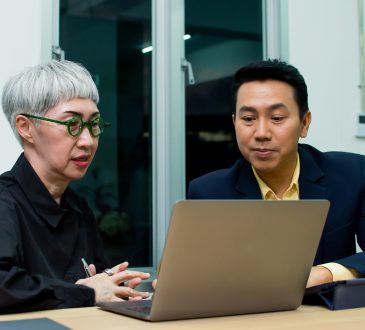 Older woman and younger male colleague talking over laptop in office