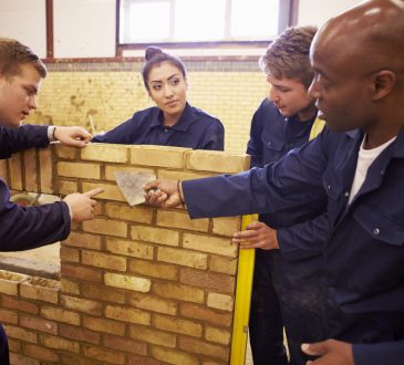 Teacher Helping Students Training To Be Builders Standing Next To Brick Wall
