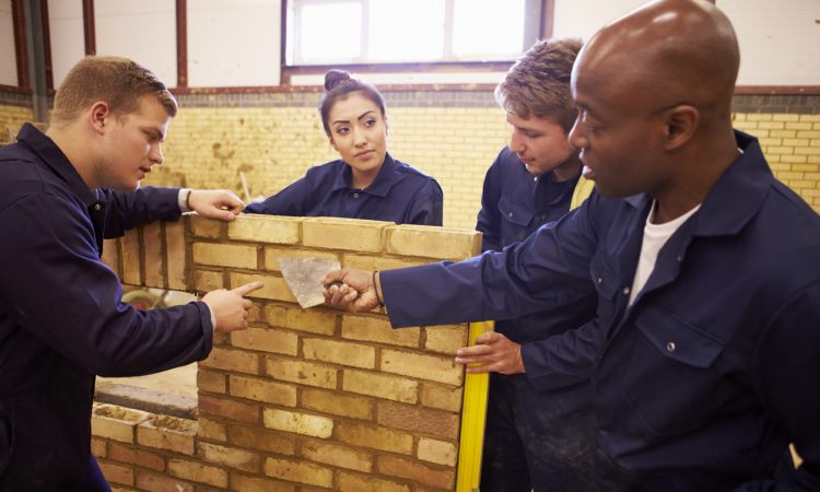 Teacher Helping Students Training To Be Builders Standing Next To Brick Wall
