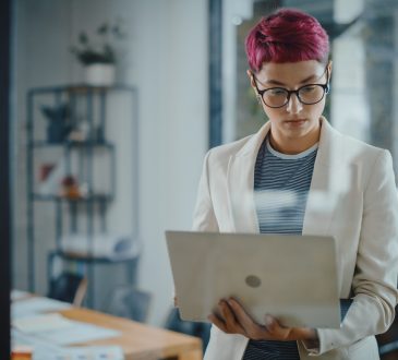 Person with short pink hair standing and holding laptop in office