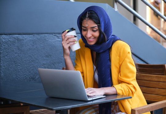 Young Indian businesswoman wearing yellow suit jacket smiling and working with a laptop at a coffee shop outdoors.