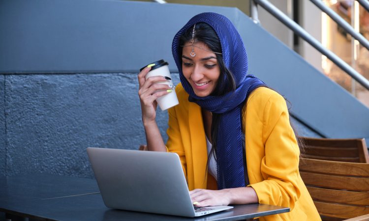 Young Indian businesswoman wearing yellow suit jacket smiling and working with a laptop at a coffee shop outdoors.
