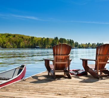 Two Muskoka chairs on a wooden dock overlooking the blue water of a lake in Muskoka, Ontario