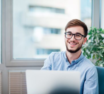 Young businessman in office working on laptop, smiling and wearing glasses