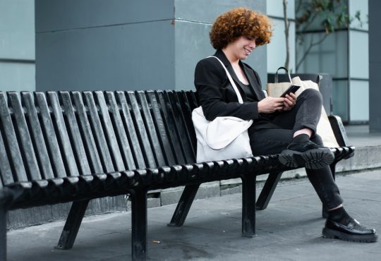Non-binary person looking at phone on black bench outside of office