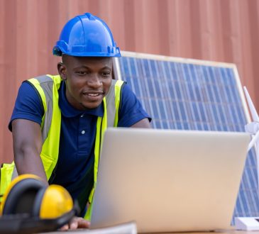 Engineer working on laptop outside in front of solar panel propped up against building
