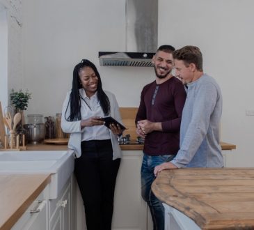 Black female real estate agent in kitchen showing gay couple around new house.