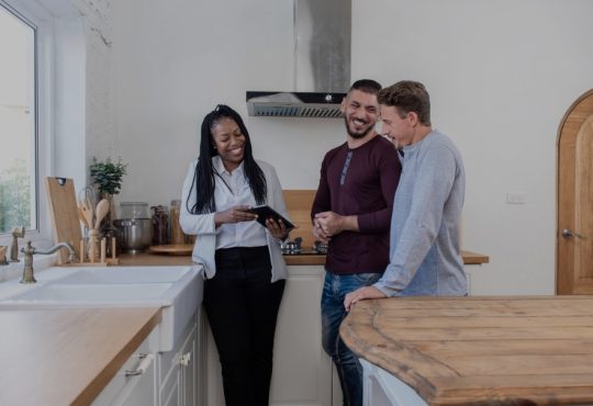 Black female real estate agent in kitchen showing gay couple around new house.