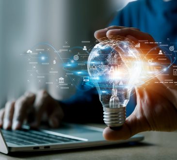 Person typing on laptop and holding glowing lightbulb in one hand with technology icons around it