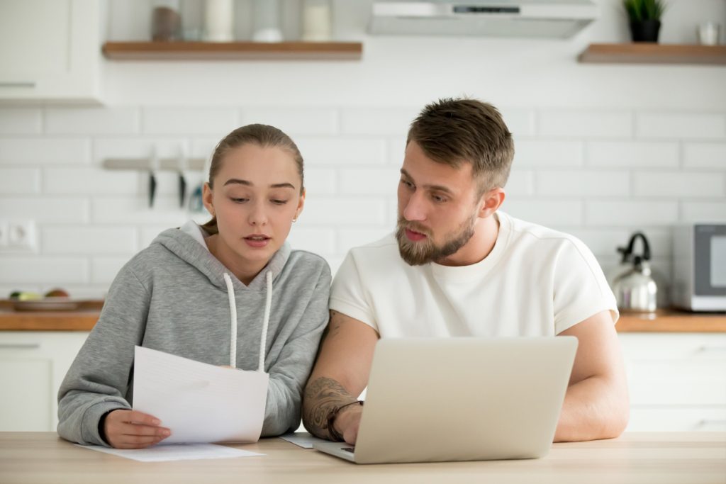 Man and woman looking at laptop and paper statement in kitchen