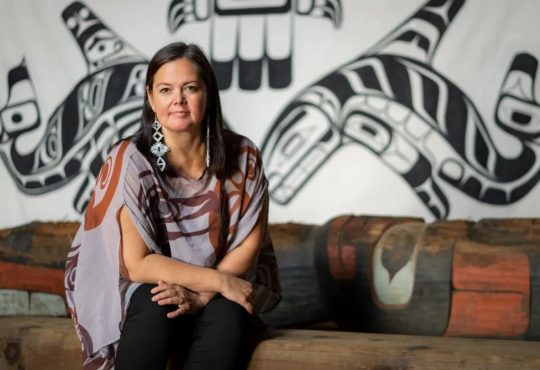 Woman sitting in front of Indigenous artwork on wall.