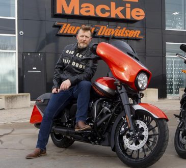 Man wearing leather jacket and blue jeans sitting on red Harley Davidson looking into camera