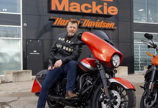 Man wearing leather jacket and blue jeans sitting on red Harley Davidson looking into camera