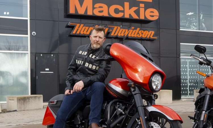 Man wearing leather jacket and blue jeans sitting on red Harley Davidson looking into camera