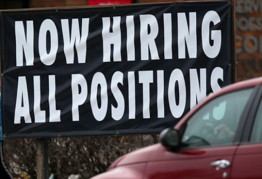 Car driving past large sign reading "NOW HIRING ALL POSITIONS"