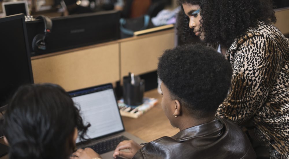 Three co-workers of varying genders working on a laptop, seen from back. The person on the right, whose face is most visible, is wearing teal eyeshadow. They are Black and have shoulder-length, curly brown hair. They are wearing a leopard-print blazer.