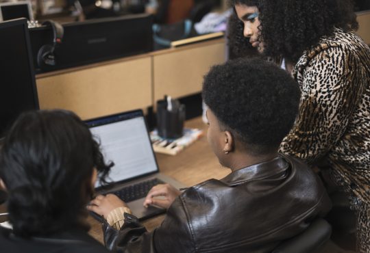 Three co-workers of varying genders working on a laptop, seen from back. The person on the right, whose face is most visible, is wearing teal eyeshadow. They are Black and have shoulder-length, curly brown hair. They are wearing a leopard-print blazer.