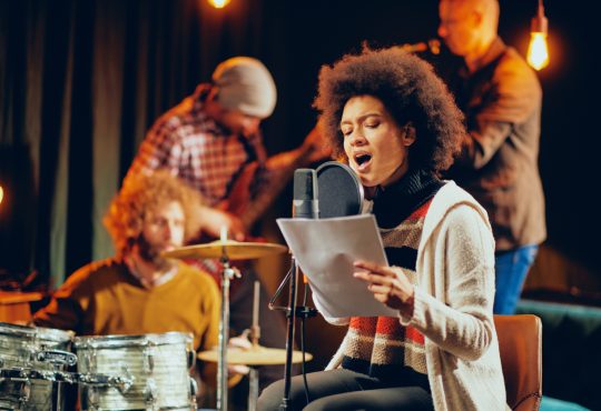 Woman with brown afro hold paper and sings, with band laying in background