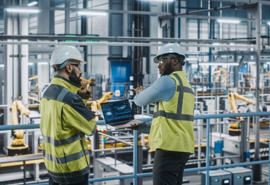 Two Car Factory Specialists in High Visibility Vests Using Laptop Computer.