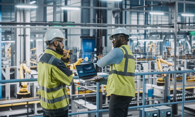 Two Car Factory Specialists in High Visibility Vests Using Laptop Computer.