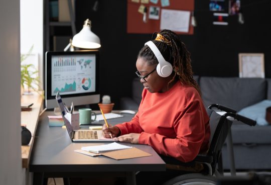 A Black woman with hair in braids in high ponytail, wearing white over-ear headphones and red long-sleeved shirt, sits in a wheelchair at a desk, taking notes in front of a laptop.