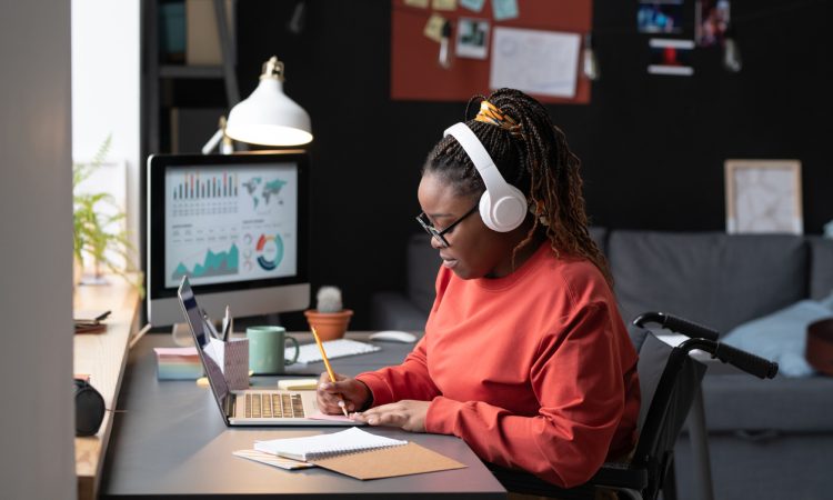 A Black woman with hair in braids in high ponytail, wearing white over-ear headphones and red long-sleeved shirt, sits in a wheelchair at a desk, taking notes in front of a laptop.