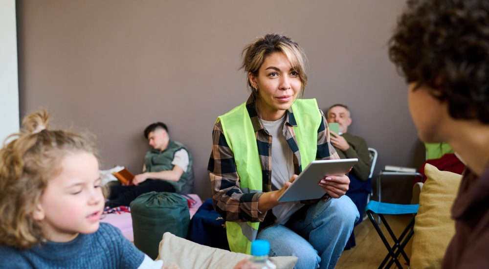 Young female volunteer wearing yellow vest squats in front of woman with son in community centre and enters their information on a tablet.