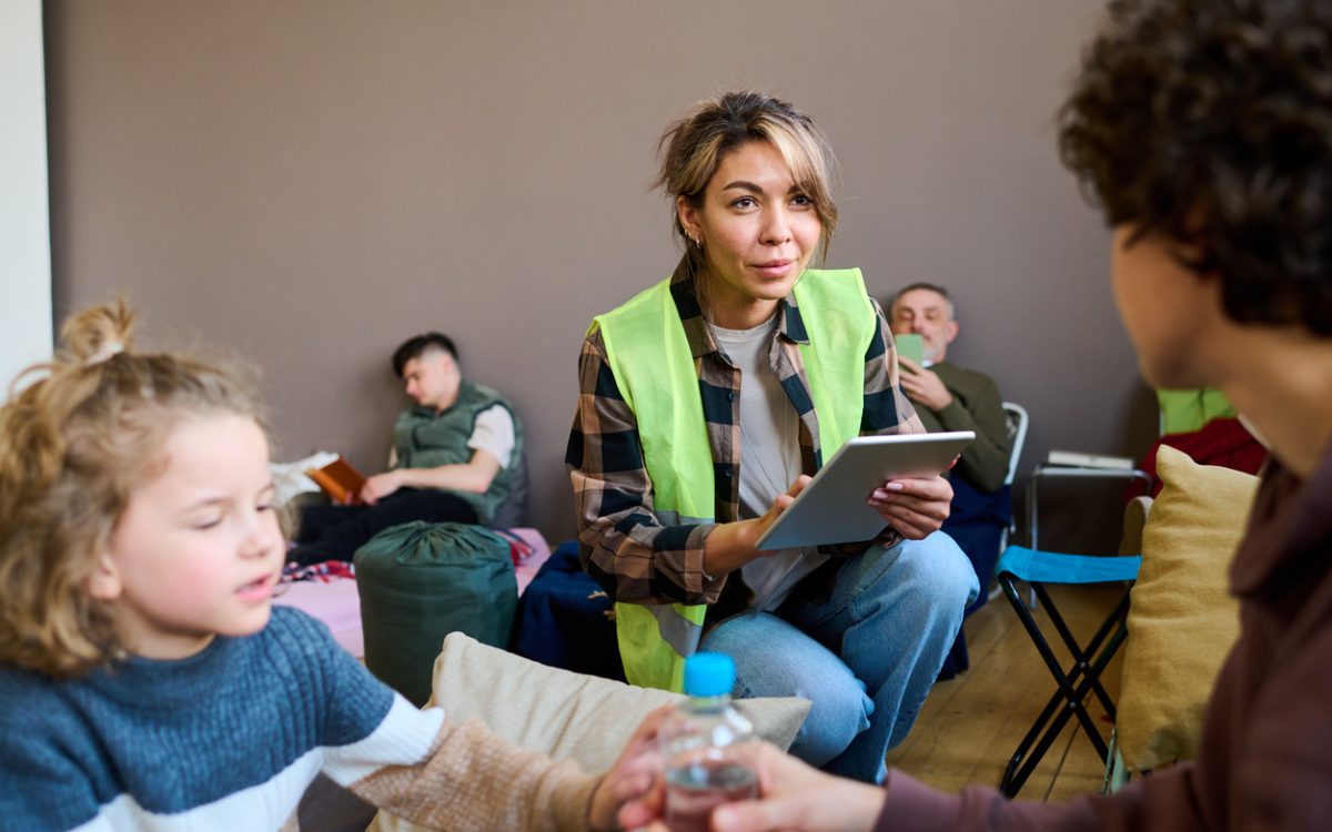 Young female volunteer wearing yellow vest squats in front of woman with son in community centre and enters their information on a tablet.