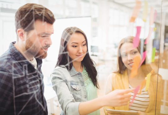 Three co-workers putting sticky notes on clear wall in office