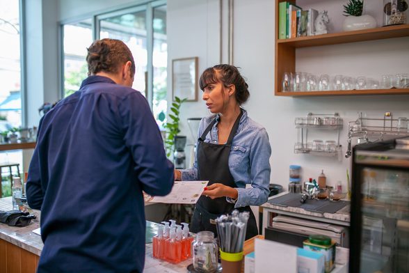 A barista wearing a blue long-sleeved shirt and a black apron takes an order from a man standing at counter wearing a long-sleeved, dark blue button-up shirt
