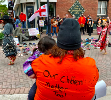 Group of people attend Truth and Reconciliation event. Person in foreground is wearing orange shirt with "Our children matter too!" handwritten on the back.