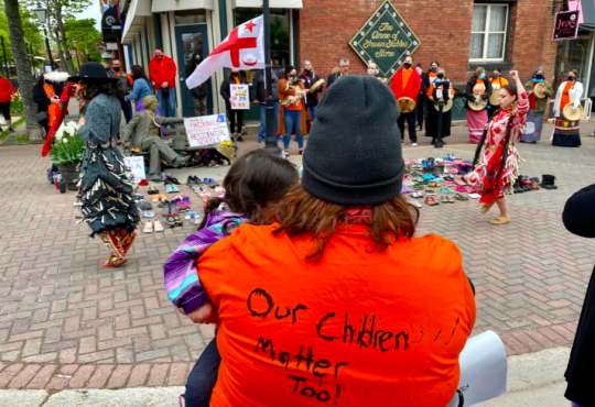 Group of people attend Truth and Reconciliation event. Person in foreground is wearing orange shirt with "Our children matter too!" handwritten on the back.