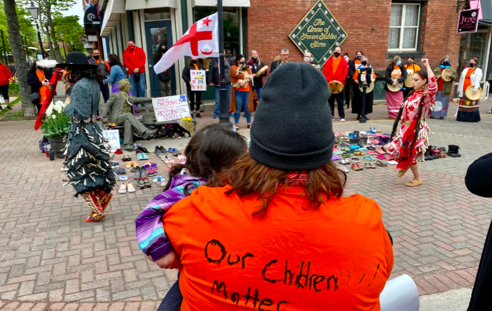 Group of people attend Truth and Reconciliation event. Person in foreground is wearing orange shirt with "Our children matter too!" handwritten on the back.