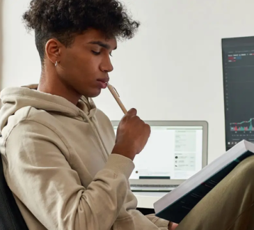 Young man sits in computer chair with textbook open in lap, holding pen to his mouth. He is wearing a beige hoodie, forest green slacks and has brown curly hair