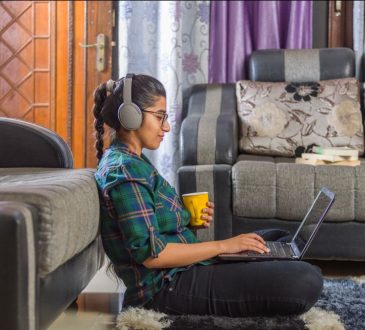 Young woman sitting on floor leaning up against couch, working on laptop in her lap while holding coffee mug in hand