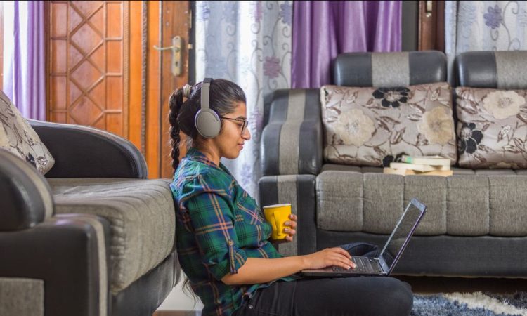 Young woman sitting on floor leaning up against couch, working on laptop in her lap while holding coffee mug in hand