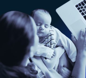 Black-and-white overhead image of woman holding infant while working on laptop