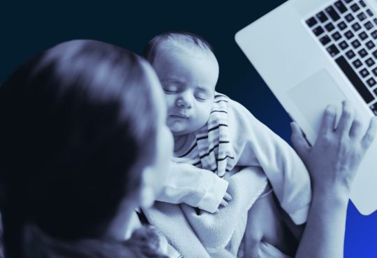 Black-and-white overhead image of woman holding infant while working on laptop