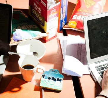 Two people working on laptops on messy desk strewn with cups and food