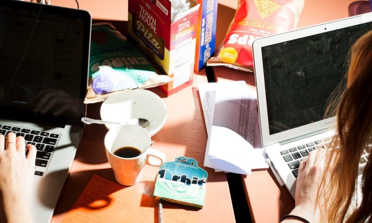 Two people working on laptops on messy desk strewn with cups and food