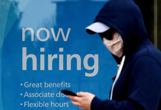 Masked person walking past business window with "now hiring" sign