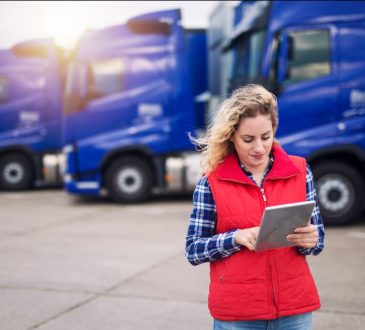 Woman standing in lot of transport trucks looking at tablet device.