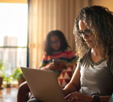 Woman with shoulder-length brown curly hair, wearing gray tshirt, sits on couch looking at laptop in her lap