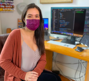 Portrait of woman wearing mask sitting at desk, facing camera. The computer screen behind her is covered in code.