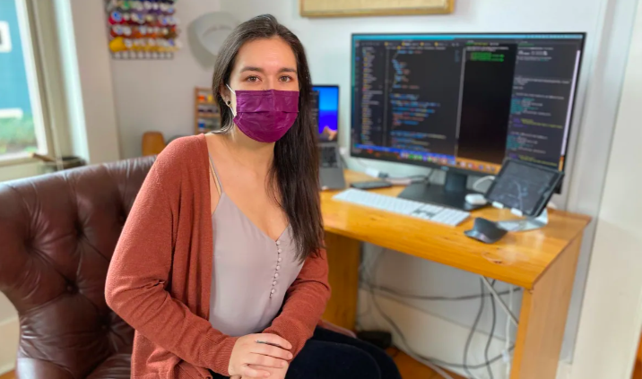 Portrait of woman wearing mask sitting at desk, facing camera. The computer screen behind her is covered in code.